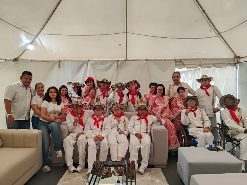 Nuestros artistas con vestuario blanco, pañoleta roja y sombrero vueltiao, sentados junto a sus profesores bajo una carpa